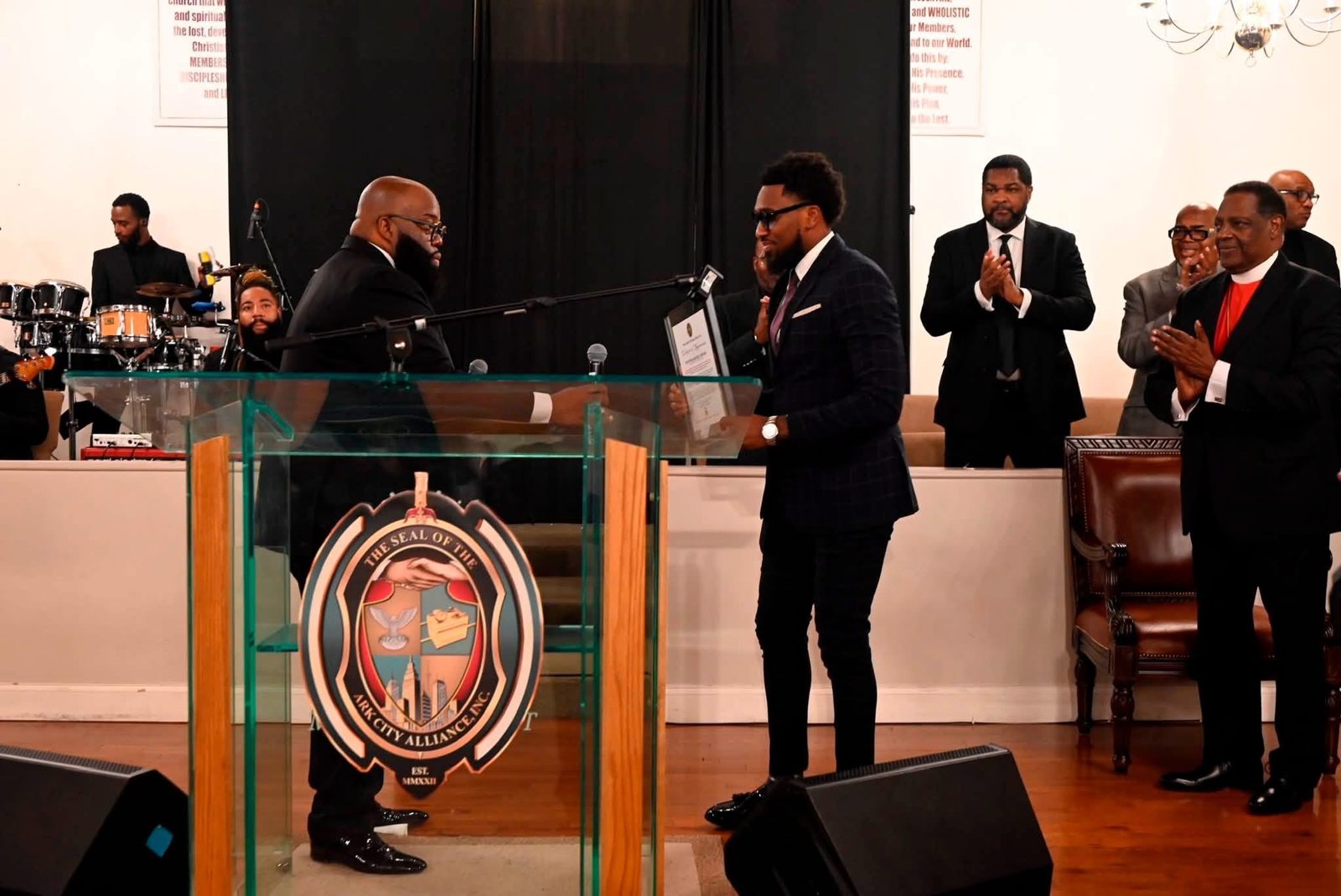 Pastor Jahleel Spence receiving a framed honor on stage from Ark City Alliance as clergy and guests applaud